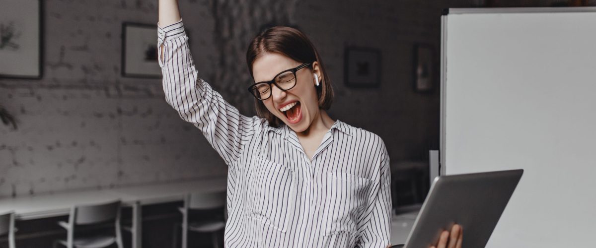 Business woman with laptop in hand is happy with success. Portrait of woman in glasses and striped blouse enthusiastically screaming and making winning gesture